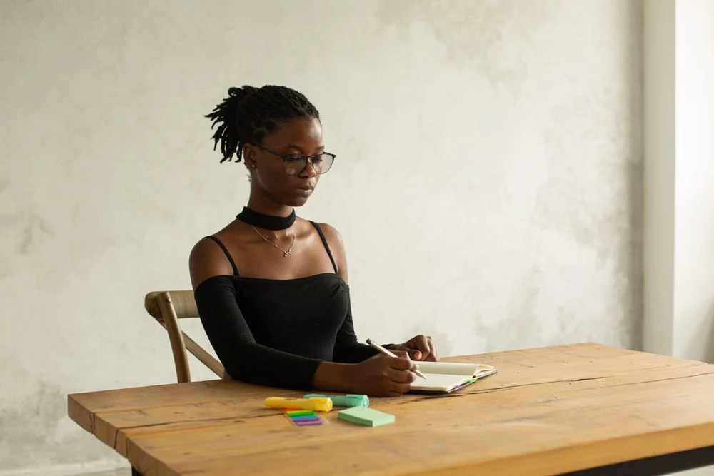 A person developing ideas in a notebook at a wooden table by a window.